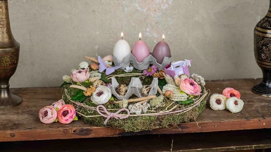 Ein Frühlingsgesteck mit rosa und weißen Blüten, Moos und drei brennenden Kerzen in Eiform